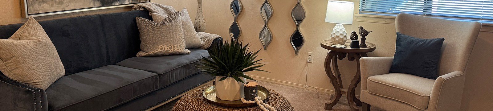 A living room with a grey couch, a small table with a plant and a lamp, and a beige armchair.